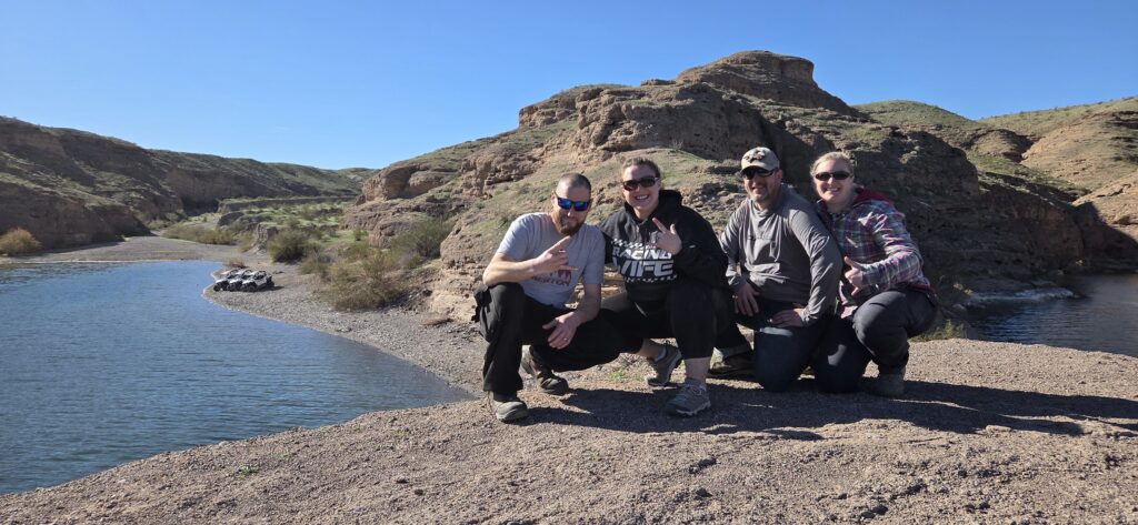 Group smiles on Colorado River bank near Las Vegas, Nevada, enjoying scenic views and outdoor adventure in stunning red hills.