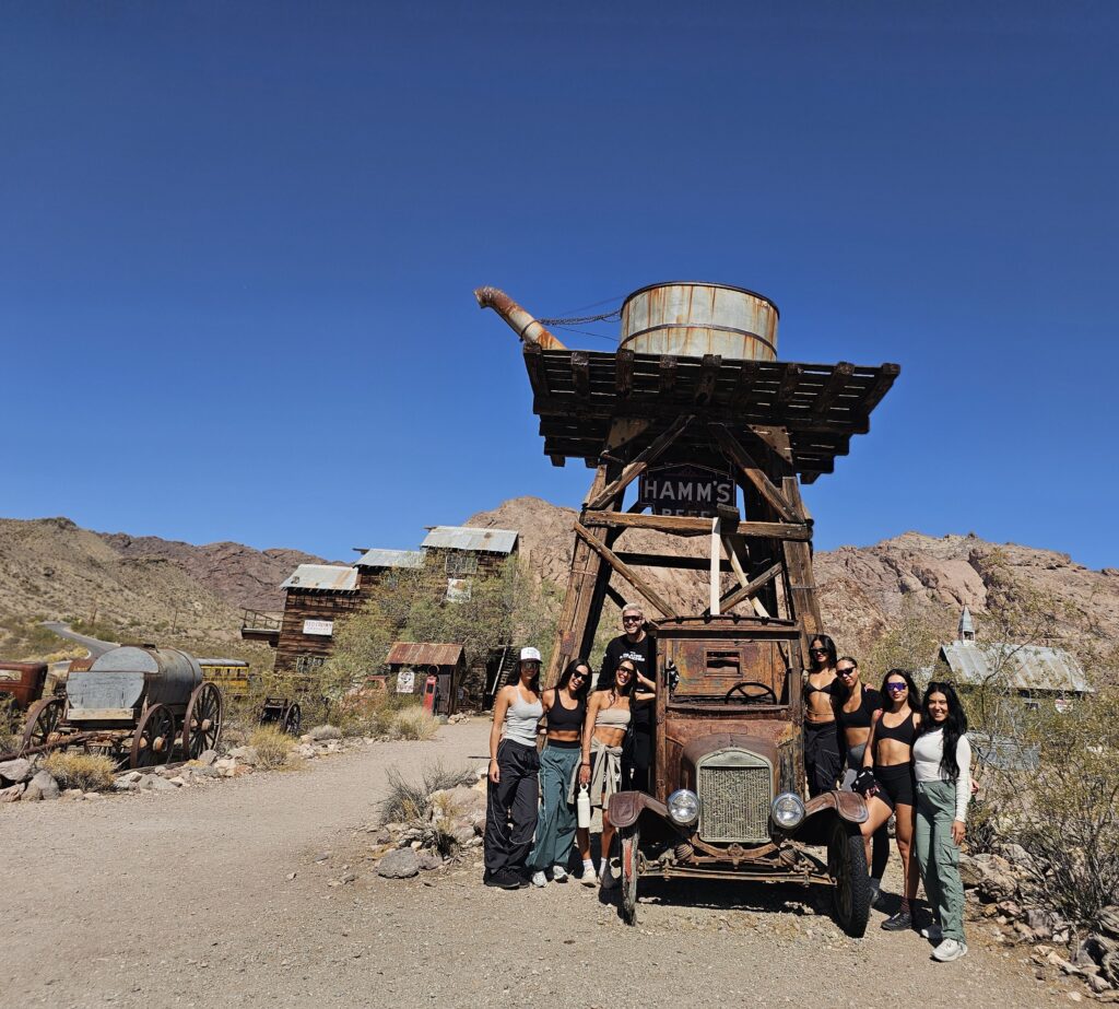 Seven visitors pose by a rusty truck and HAMMS water tower in Nelson Ghost Town, Nevada near Las Vegas—iconic sightseeing spot.
