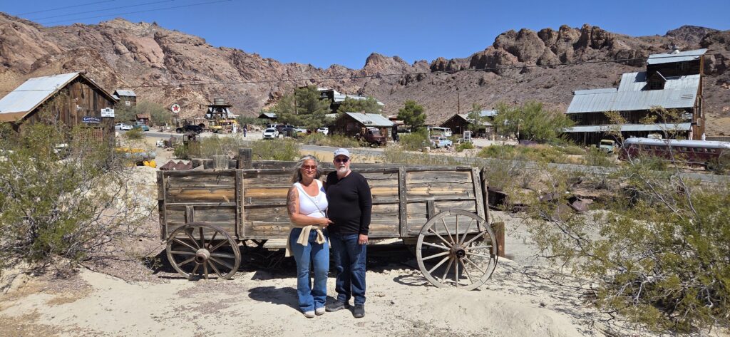 Smiling couple by old wagon in Eldorado Canyon near Las Vegas, Nevada, with ghost town sights and desert mountains in the background.