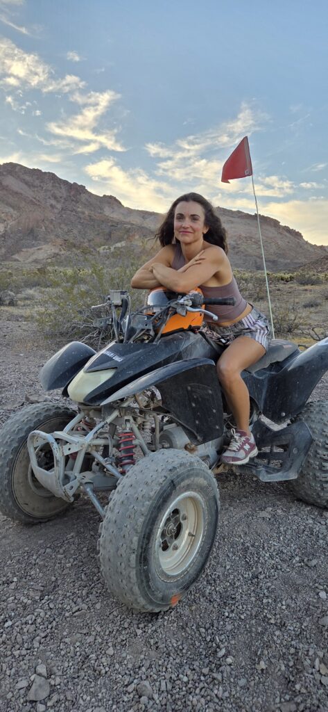 Woman on Las Vegas ATV tour rides black RZR through Nevada desert near mountains, red flag flying under partly cloudy sky.