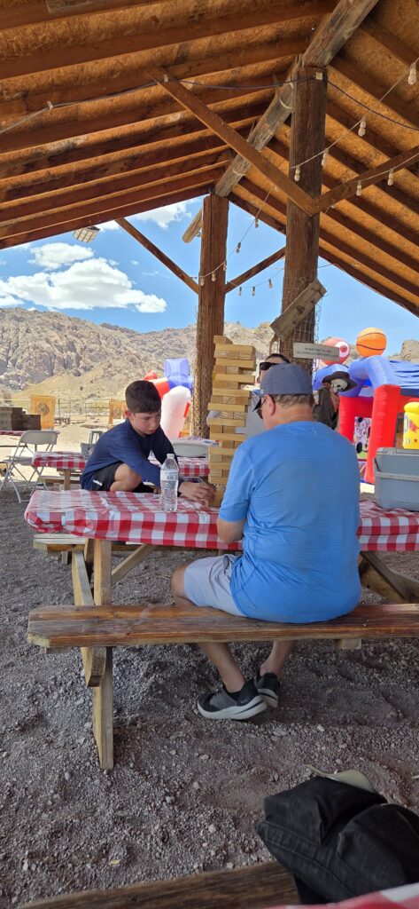 Two people play giant Jenga at a picnic in Las Vegas, Nevada, with mountains and inflatable structures in the background.