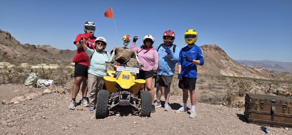 Group in helmets and outdoor gear pose by yellow ATV on Nevada desert tour near Las Vegas, treasure chest nearby, mountains behind.