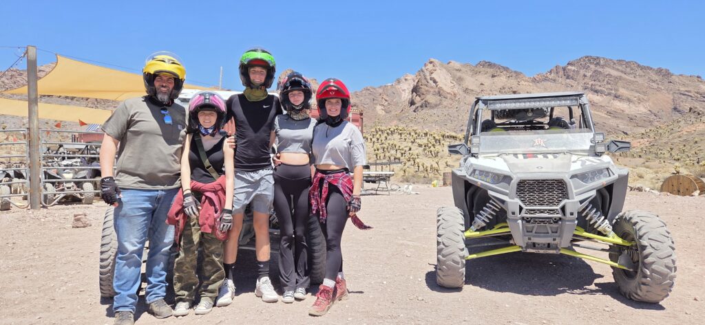 Group in helmets and casual gear pose by ATV near Las Vegas with Nevada mountains, blue sky—ideal for RZR off-road tours.