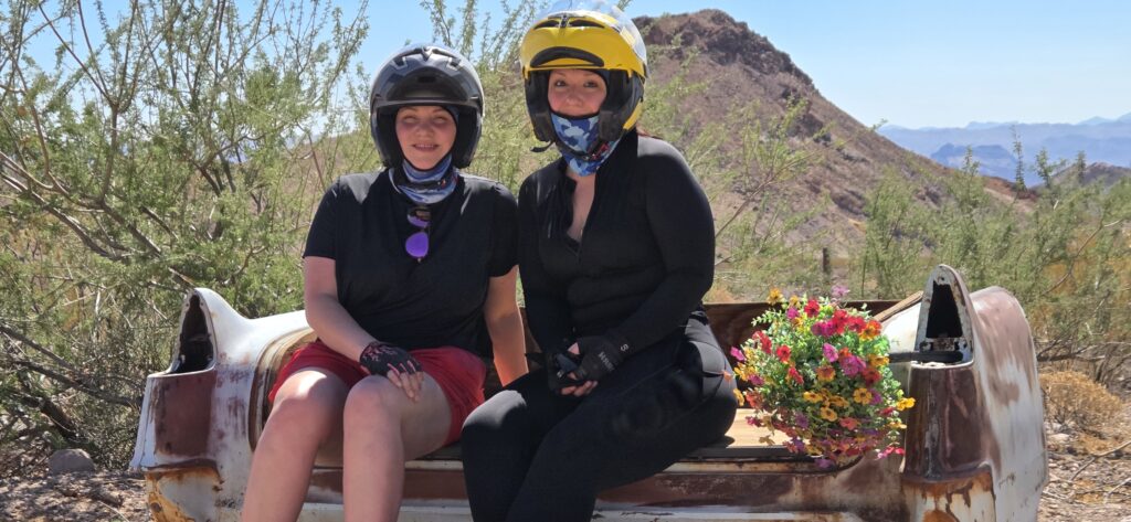 Two adventurers in helmets and gloves relax on a car trunk in the Nevada desert near Las Vegas, flowers beside them, mountains behind.