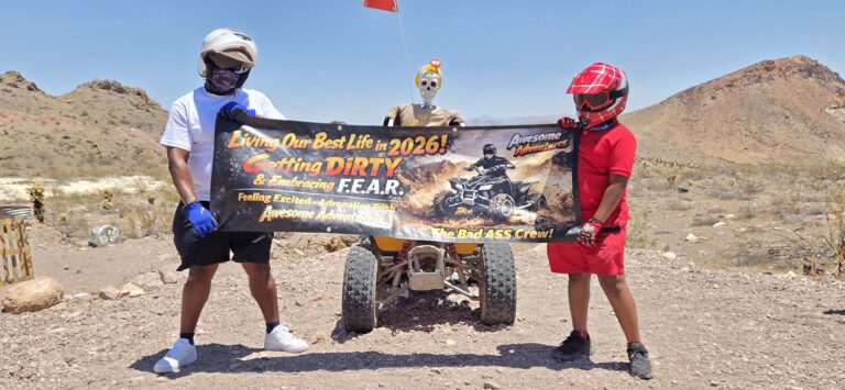 Two in helmets hold a banner for 2026 ATV tours near Las Vegas; skeleton on ATV, Nevada desert hills and dry terrain behind.