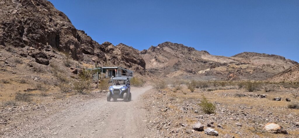 A blue off-road RZR drives a dusty Nevada desert trail near Las Vegas, with mountains and a Ghost Town under clear blue skies.