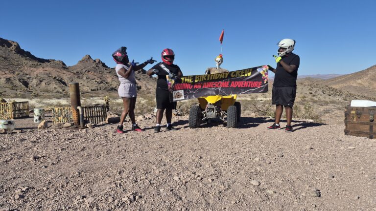 Group in helmets by a yellow ATV on a Nevada desert tour, holding “The Birthday Crew” banner; rocky hills and blue sky behind.