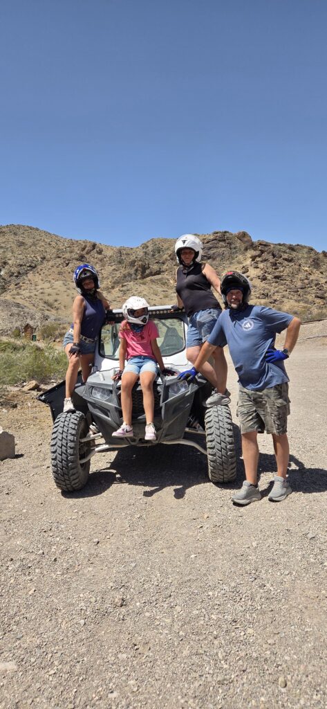 Group enjoys Las Vegas ATV tour in Nevada desert; child on RZR hood, adults smiling, clear blue sky, rocky terrain in background.