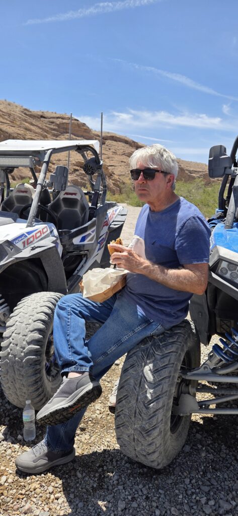 Man in sunglasses enjoys a sandwich on an ATV tire during a Nevada RZR off-road tour near Las Vegas desert terrain.