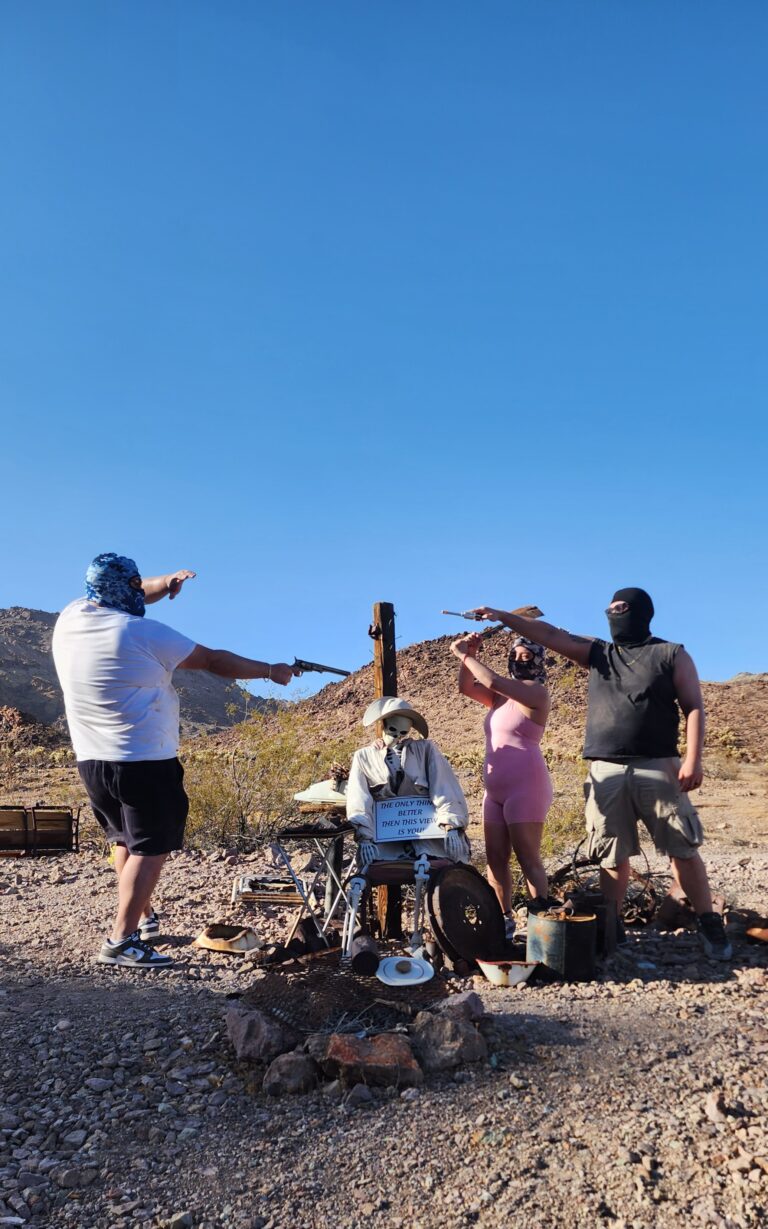 In the Nevada desert near Las Vegas, three masked figures aim guns at a costumed mannequin, with mountains and blue sky behind.