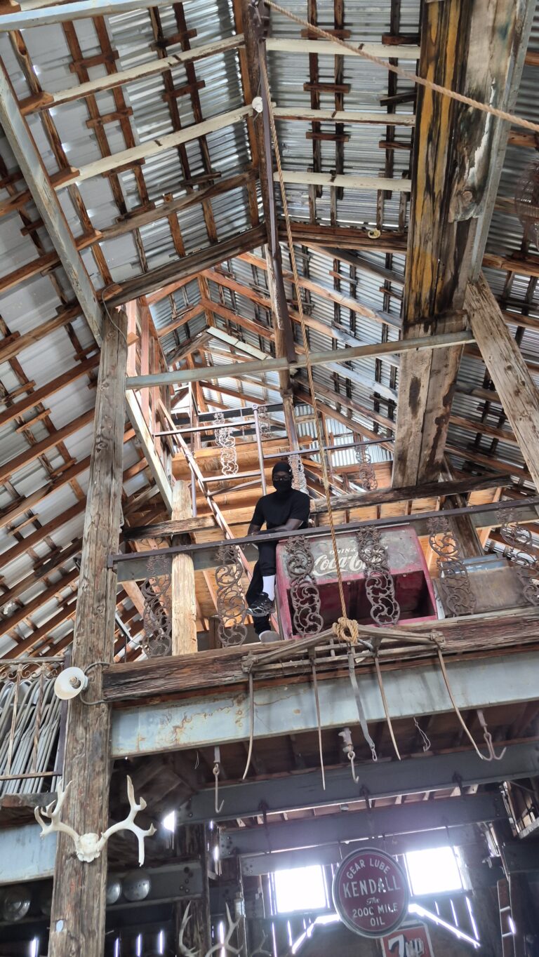 A person in black on a rustic barn balcony near the Colorado River in Nevada; chains, antlers, and Kendall sign add ghost town vibes.