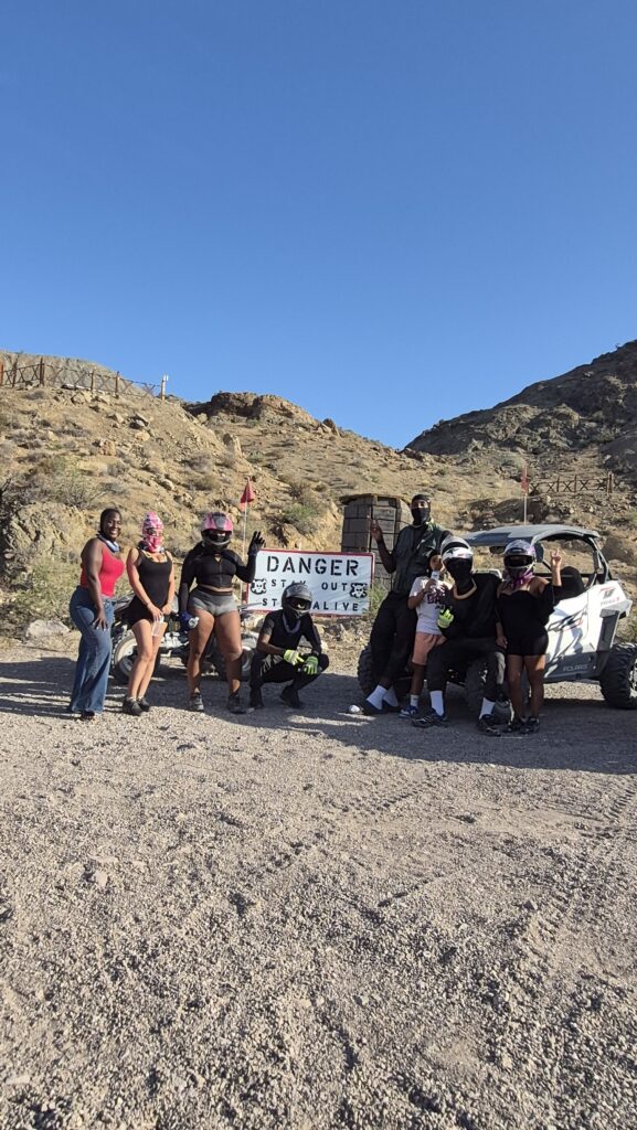 Adventurers in helmets pose by a "Danger: Stay Out, Stay Alive" sign and white ATV on a Nevada desert ATV tour near Las Vegas.