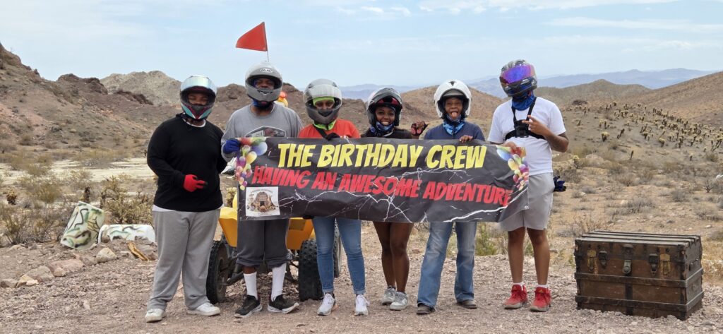 Group in helmets and casual wear holds a Birthday Crew Adventure banner during an ATV tour near Nevada’s rocky desert hills by Las Vegas.