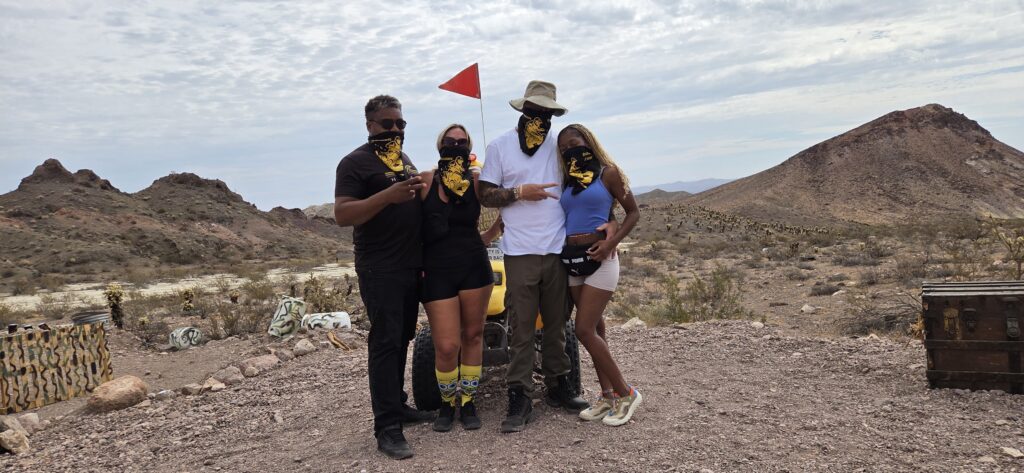 Four adventurers in yellow-and-black bandanas pose in a Nevada desert near Las Vegas with treasure chests and red flag, perfect for ATV tours.
