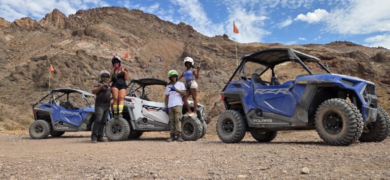 Group in helmets and outdoor gear pose by Polaris RZR vehicles on rocky Nevada terrain during an ATV tour near Las Vegas.