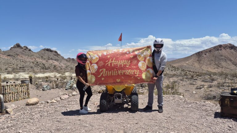 Two people in helmets hold a Happy Anniversary banner over an ATV in the Nevada desert near Las Vegas, with rocky hills and blue sky.