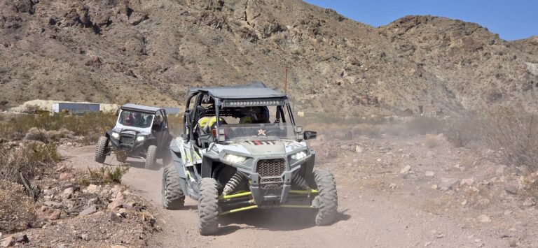 ATV riders speed along a dusty desert trail near Las Vegas, Nevada, with rocky hills and dry bushes under a clear blue sky.