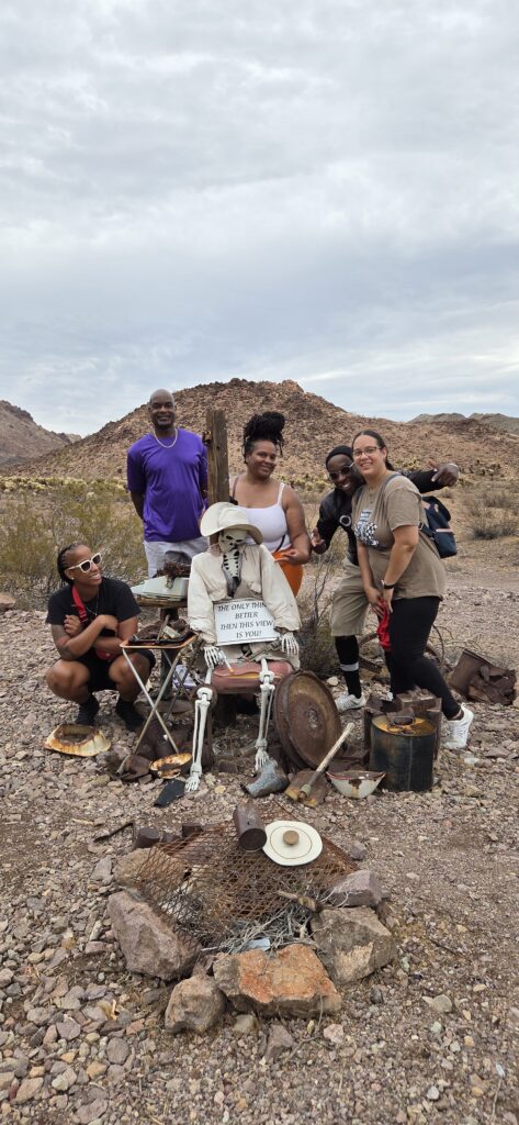 Group poses in Nevada desert near skeleton with 'The Old Man' sign, rocky terrain, Las Vegas day trip sightseeing backdrop.