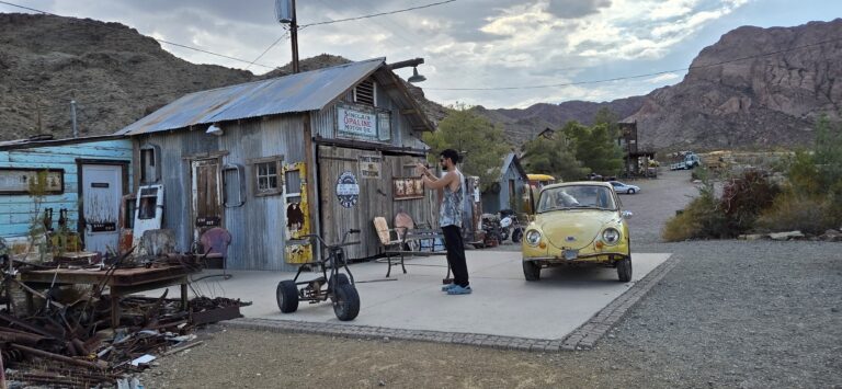 A visitor photographs a rustic building and vintage yellow car in a Nevada ghost town near the Colorado River, with mountains behind.
