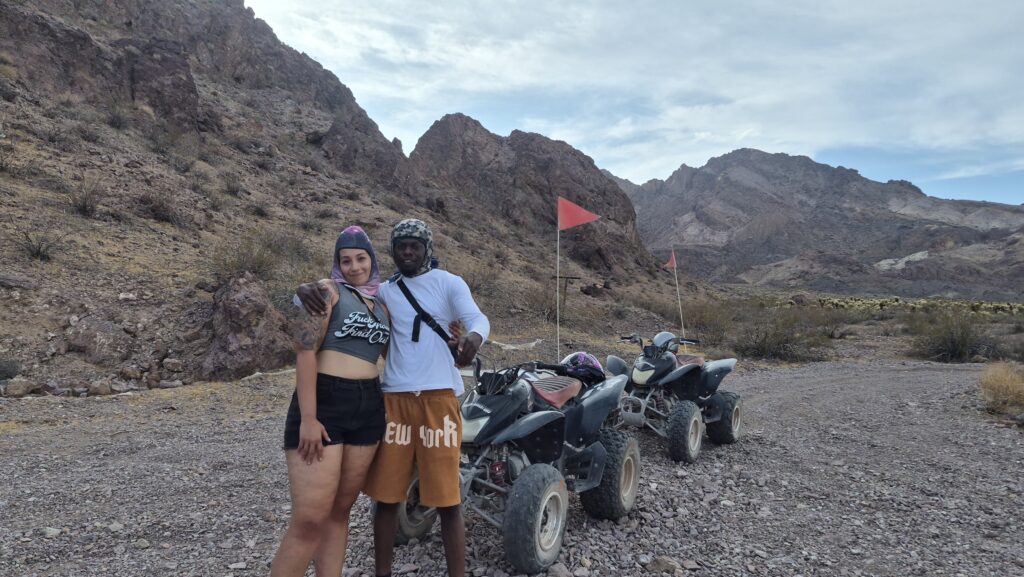 Two people in helmets stand on a Nevada desert trail with ATVs near mountains, ready for an exciting Las Vegas ATV tour adventure.
