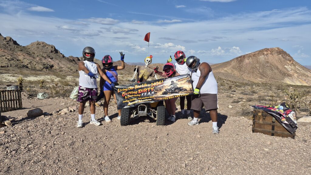 Group in helmets and gloves pose with a banner before an ATV on a Nevada desert RZR tour near Las Vegas, mountains in the background.