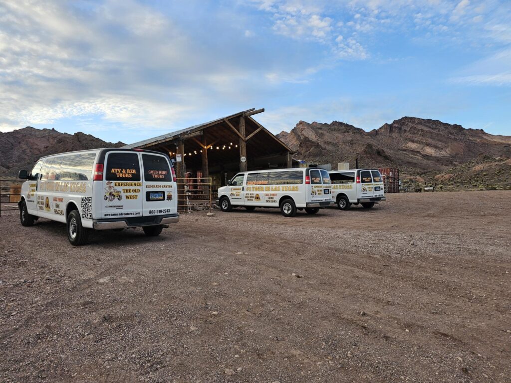 White tour vans with colorful logos await near a rustic Nevada building—ideal for Las Vegas ATV tours or Colorado River adventures.
