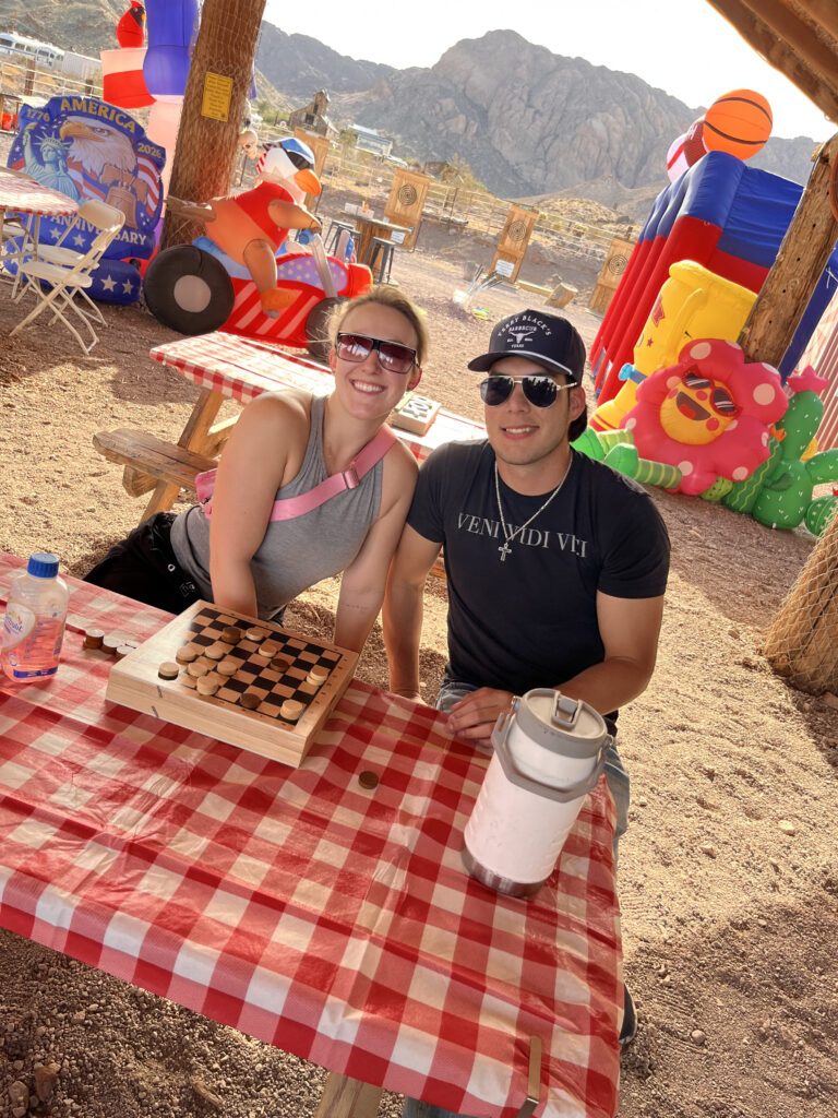 Two people enjoy a checkerboard game at a picnic table near Las Vegas, Nevada, with desert mountains and colorful inflatables nearby.