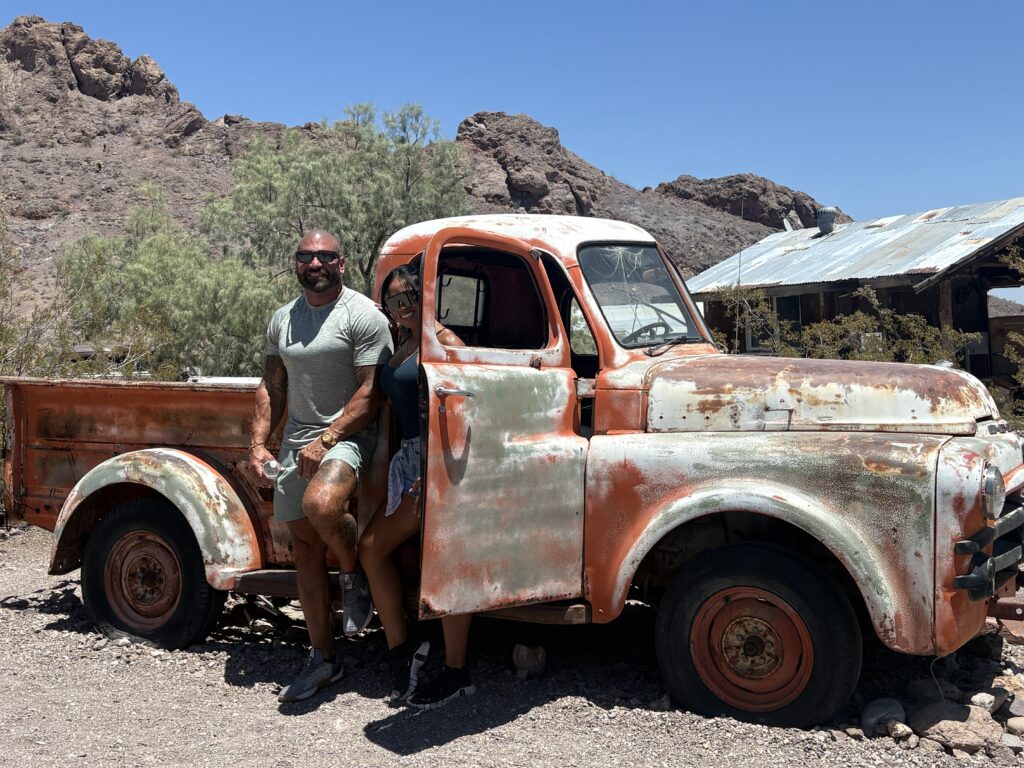 Two adventurers by a vintage orange pickup in Nevada desert, near Las Vegas Ghost Town—perfect for ATV or RZR off-road tours.