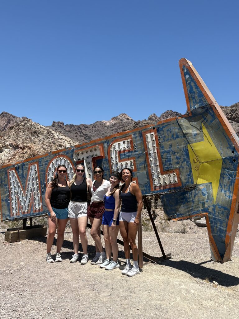 Five women in athletic wear pose by a vintage MOTEL sign near the Nevada desert, ideal for Las Vegas ATV tours and Ghost Town adventures.