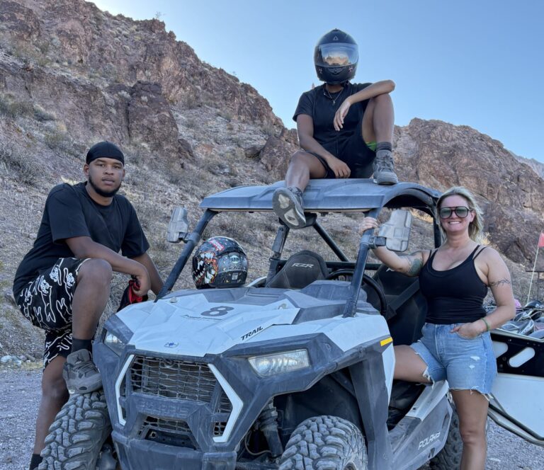 Three people enjoy a Nevada ATV tour near Las Vegas, posing with an off-road vehicle in the rocky desert by the Colorado River.