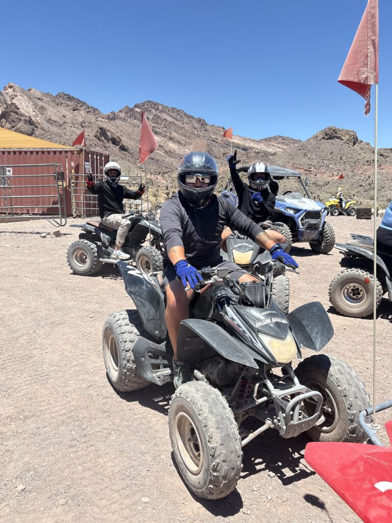 Adventurers in helmets ride ATVs near Las Vegas in the Nevada desert, posing with peace signs on an epic ATV tour under blue skies.