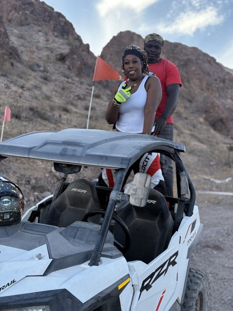 Two people pose by an RZR during a Nevada desert ATV tour near Las Vegas; orange safety flags and rocky landscape in view.