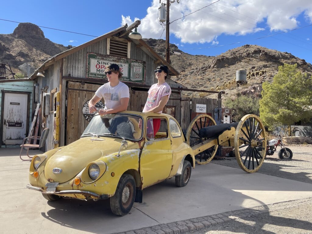 Playful duo in broken yellow car by vintage cannon at Nelson Ghost Town near Las Vegas—perfect for Nevada sightseeing adventures.
