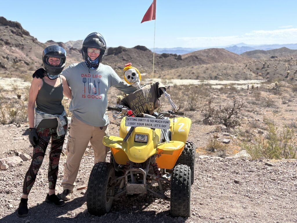 Adventurers in helmets pose by a yellow ATV with a skeleton rider near Las Vegas, Nevada, during an off-road desert tour.