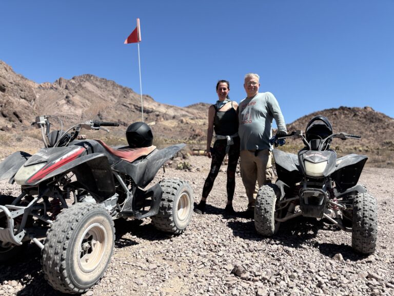 Two people on a Nevada desert ATV tour near Las Vegas, standing by RZR vehicles with mountains and a red flag in the background.