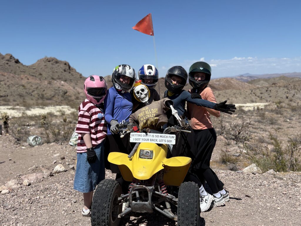 Group in helmets poses with a skeleton on a yellow ATV during a Nevada desert ATV tour near Las Vegas under blue skies.