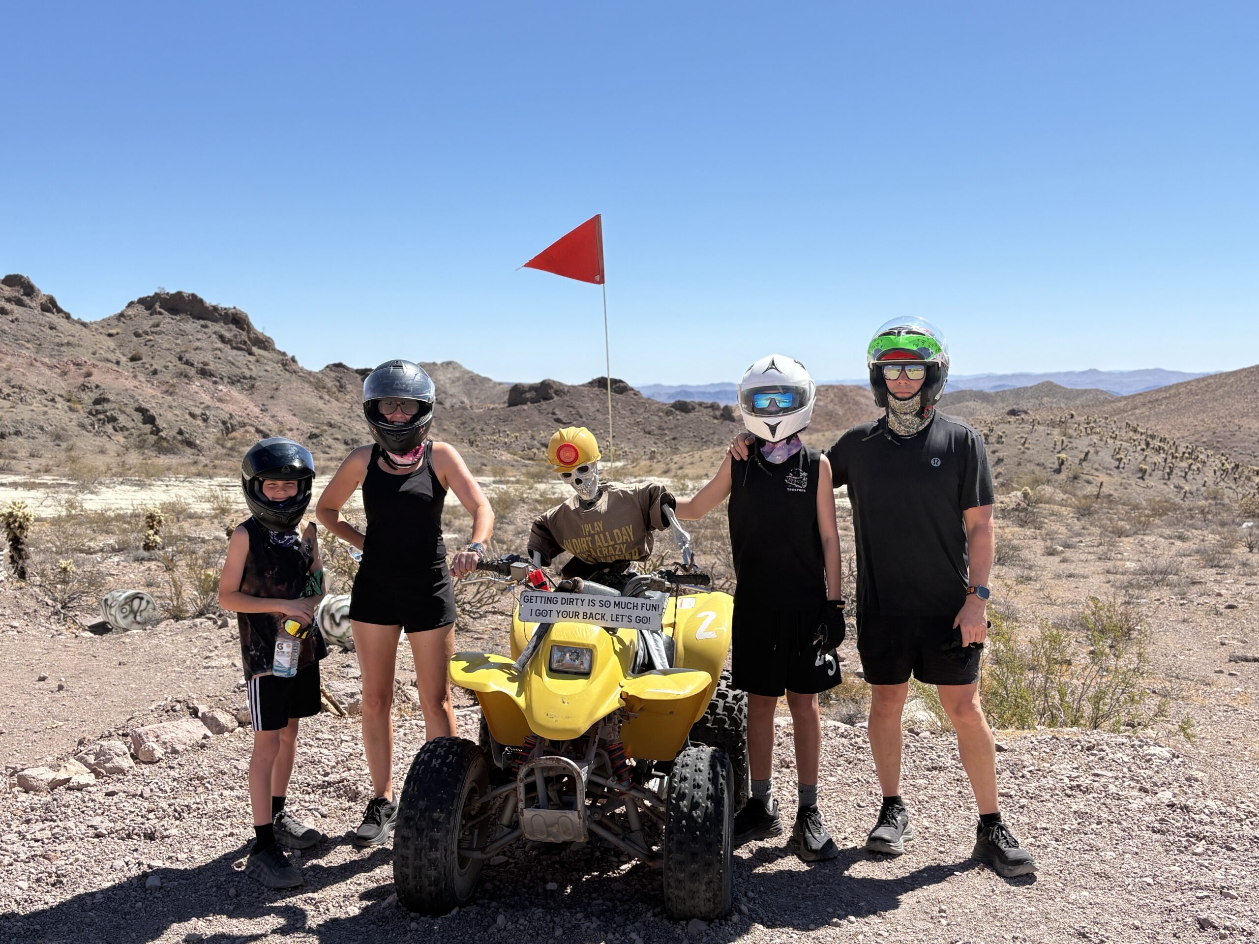 Four riders in black gear pose by a yellow ATV, scarecrow, and red flag on a Nevada desert trail near Las Vegas mountains.