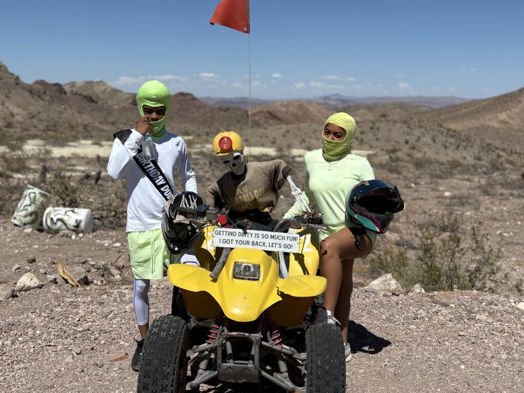 Three riders in neon face masks pose by a yellow ATV in the Nevada desert, ready for Las Vegas RZR off-road tours.