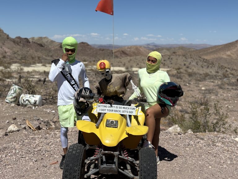 Three riders in neon face masks pose by a yellow ATV in the Nevada desert, ready for Las Vegas RZR off-road tours.