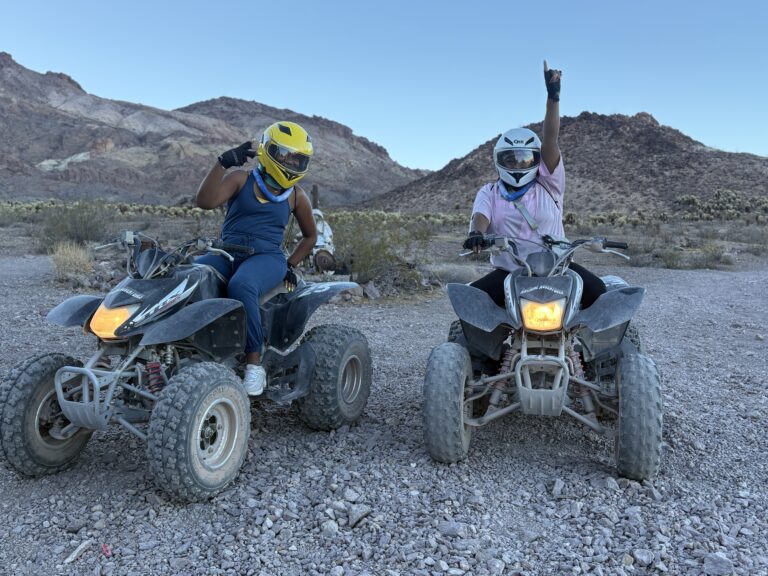 Two riders in helmets and masks enjoy an ATV tour near the Colorado River, Nevada desert, with mountains in the background.