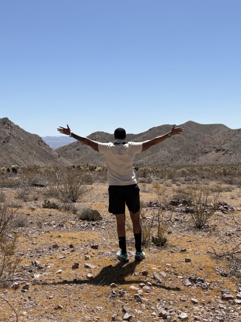 A person with arms outstretched in Nevada’s rocky desert near Las Vegas, overlooking distant mountains and the Colorado River.