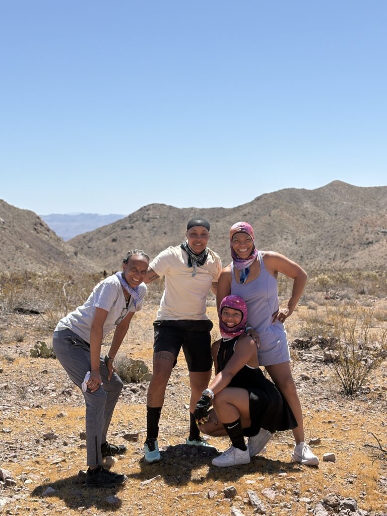 Group of four smiling hikers on a sunny Nevada desert trail near the Colorado River and Las Vegas, mountains in the background.