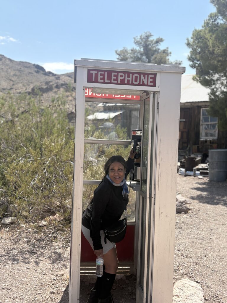 Woman in old-fashioned phone booth with water bottle at a Nevada ghost town near Las Vegas, surrounded by desert plants and rustic buildings.