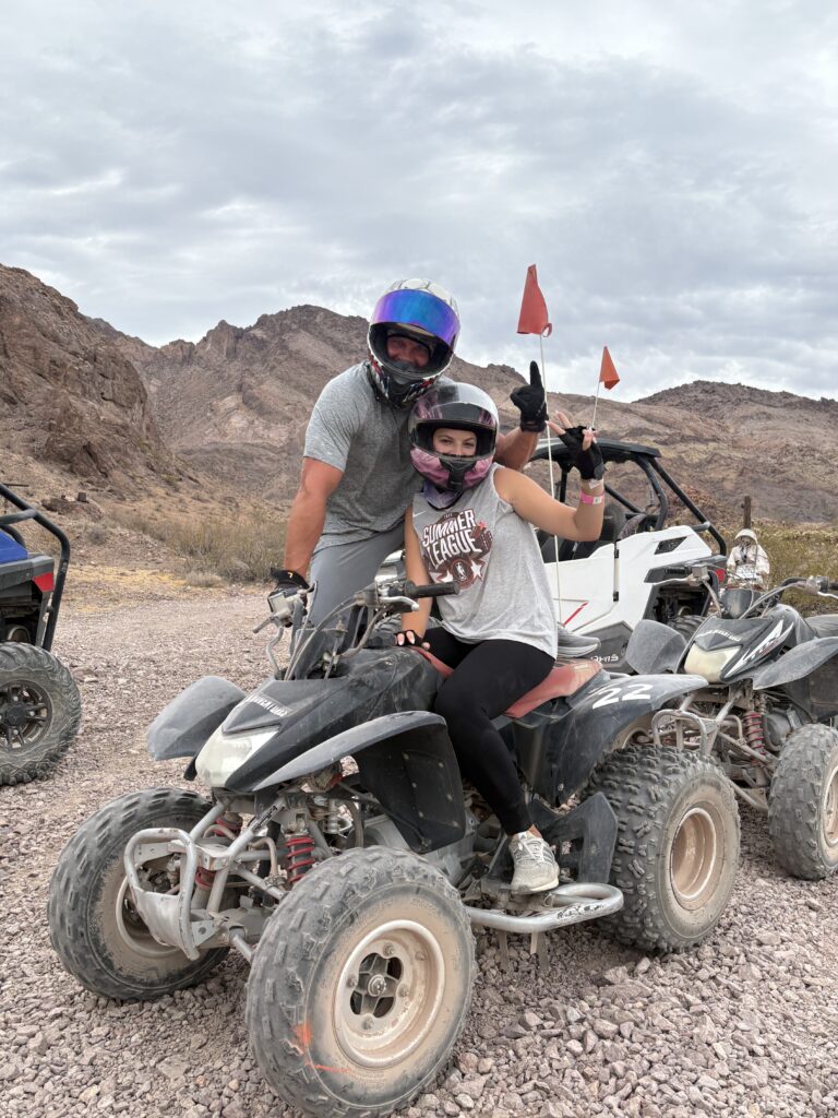 Two riders on an ATV in Nevada’s rocky desert, flashing a peace sign. Off-road vehicles and mountains near Las Vegas visible behind.