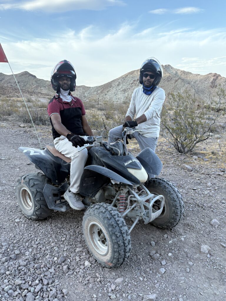Two riders in helmets and sunglasses pose with a black ATV in Nevada’s desert near Las Vegas, perfect for ATV tours and off-road fun.