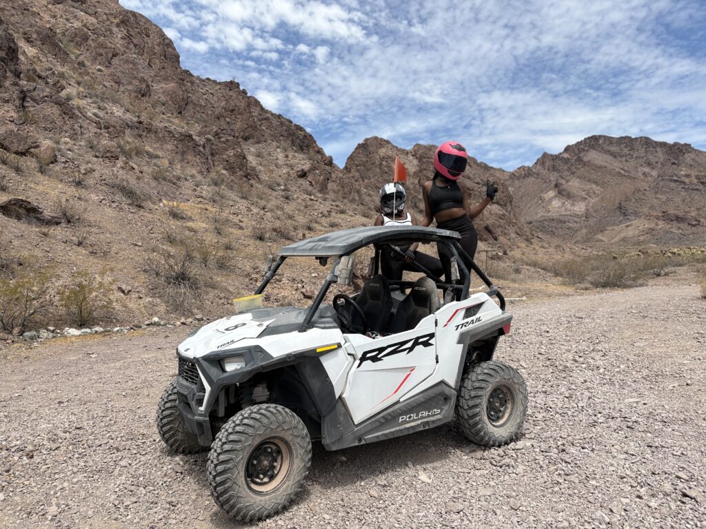 Two people in helmets stand by a white RZR on rocky Nevada terrain, perfect for Las Vegas ATV tours near the Colorado River.