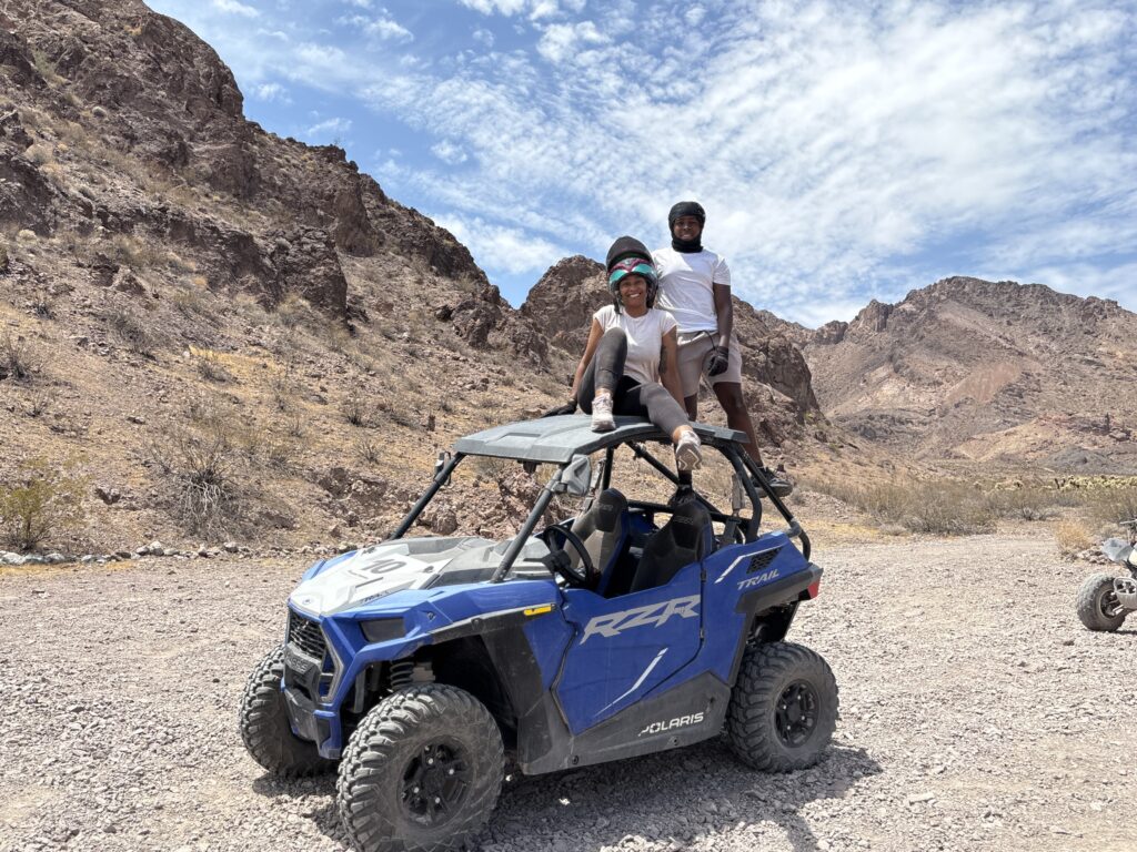 Two people in helmets on a blue Polaris RZR during an ATV tour near Las Vegas, Nevada, with desert mountains and sky behind.