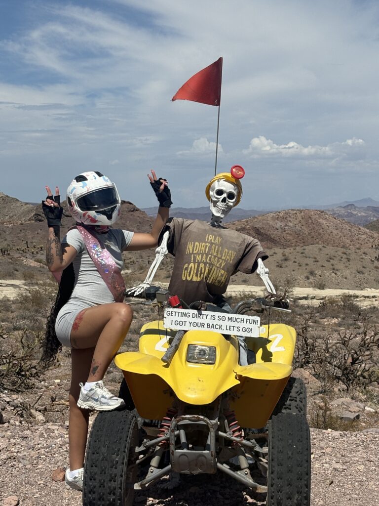 Woman in helmet on yellow ATV beside skeleton near Nevada desert hills, sign says "Getting dirty is so much fun!"—Las Vegas ATV tour.