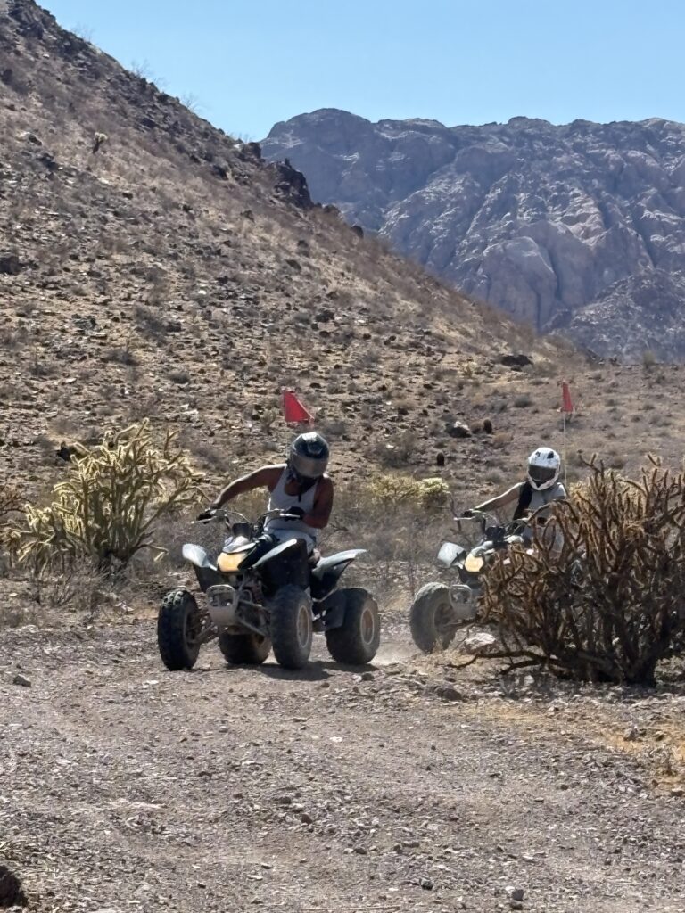 ATV riders explore a rocky desert trail near Las Vegas, Nevada, with mountains and dry bushes under clear blue skies.
