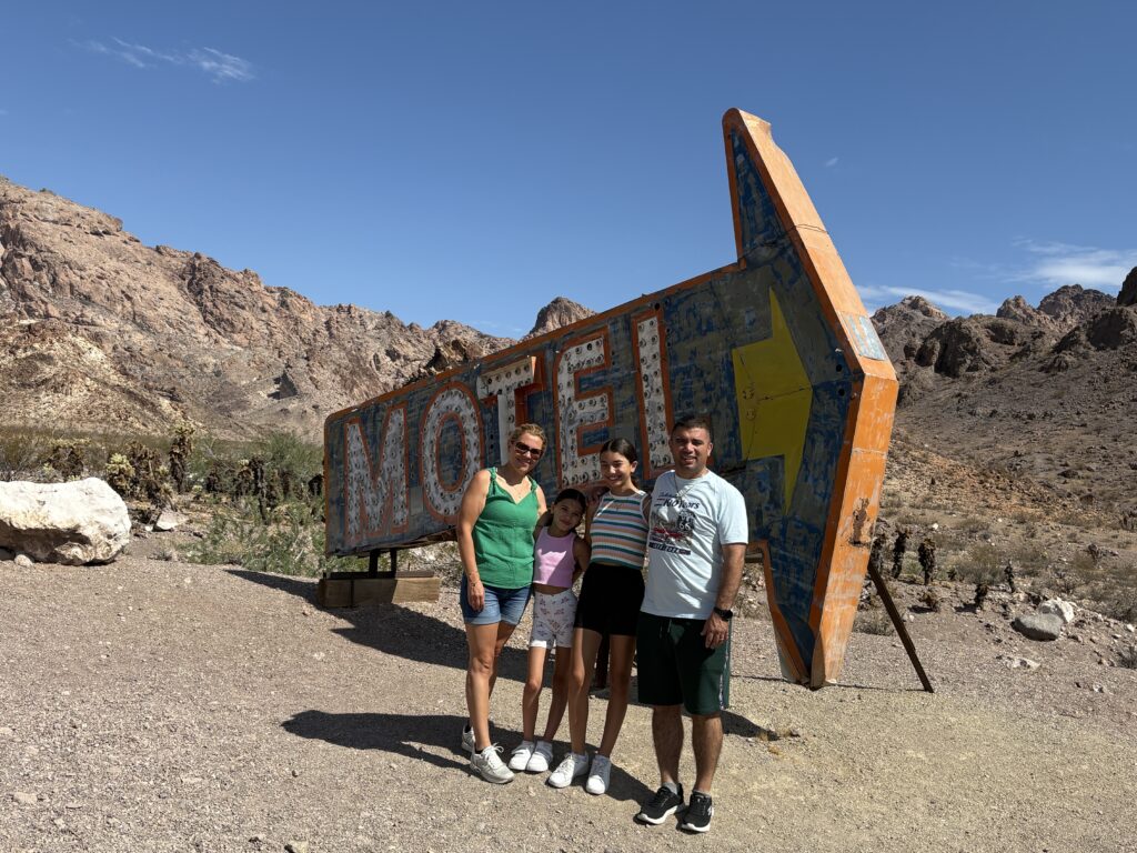 Four people pose by a vintage arrow-shaped MOTEL sign in a Nevada desert near Las Vegas, perfect for ghost town sightseeing.